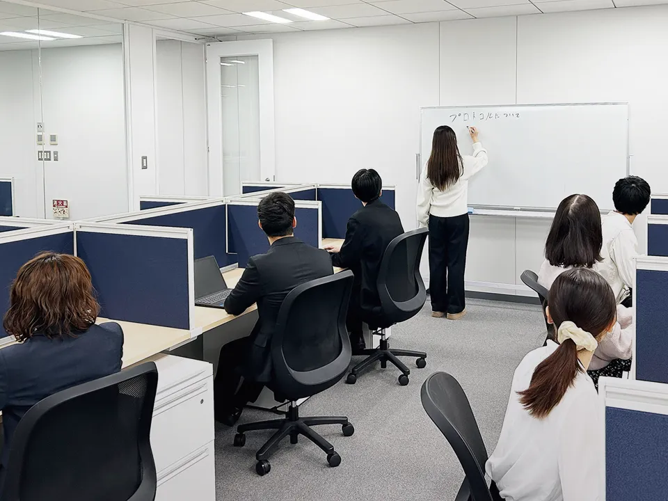 ホワイドボードに書き込んでいる女性社員と着席してそれを見ている社員たち