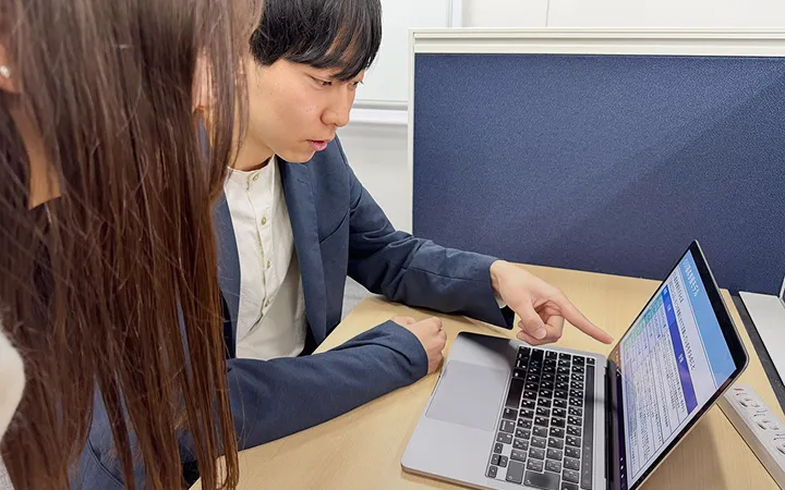 PC画面を指さして説明している男性社員とそれを聞いている女性社員