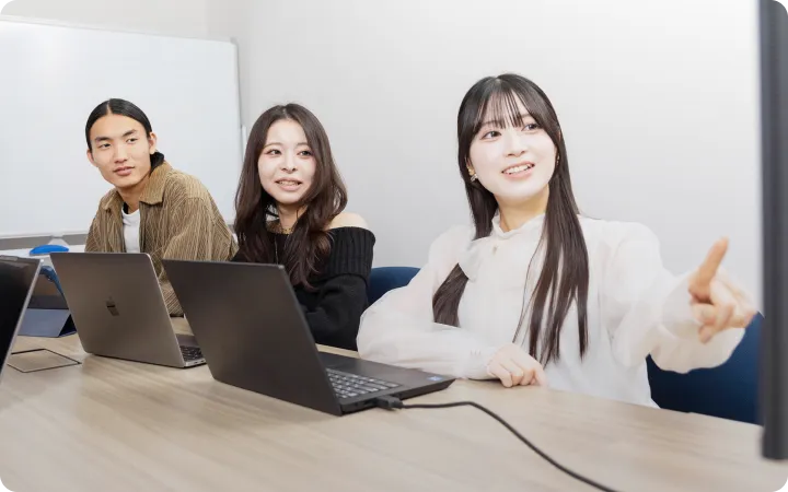 モニターを指さして説明をしている女性社員とそれを聞いている社員たち